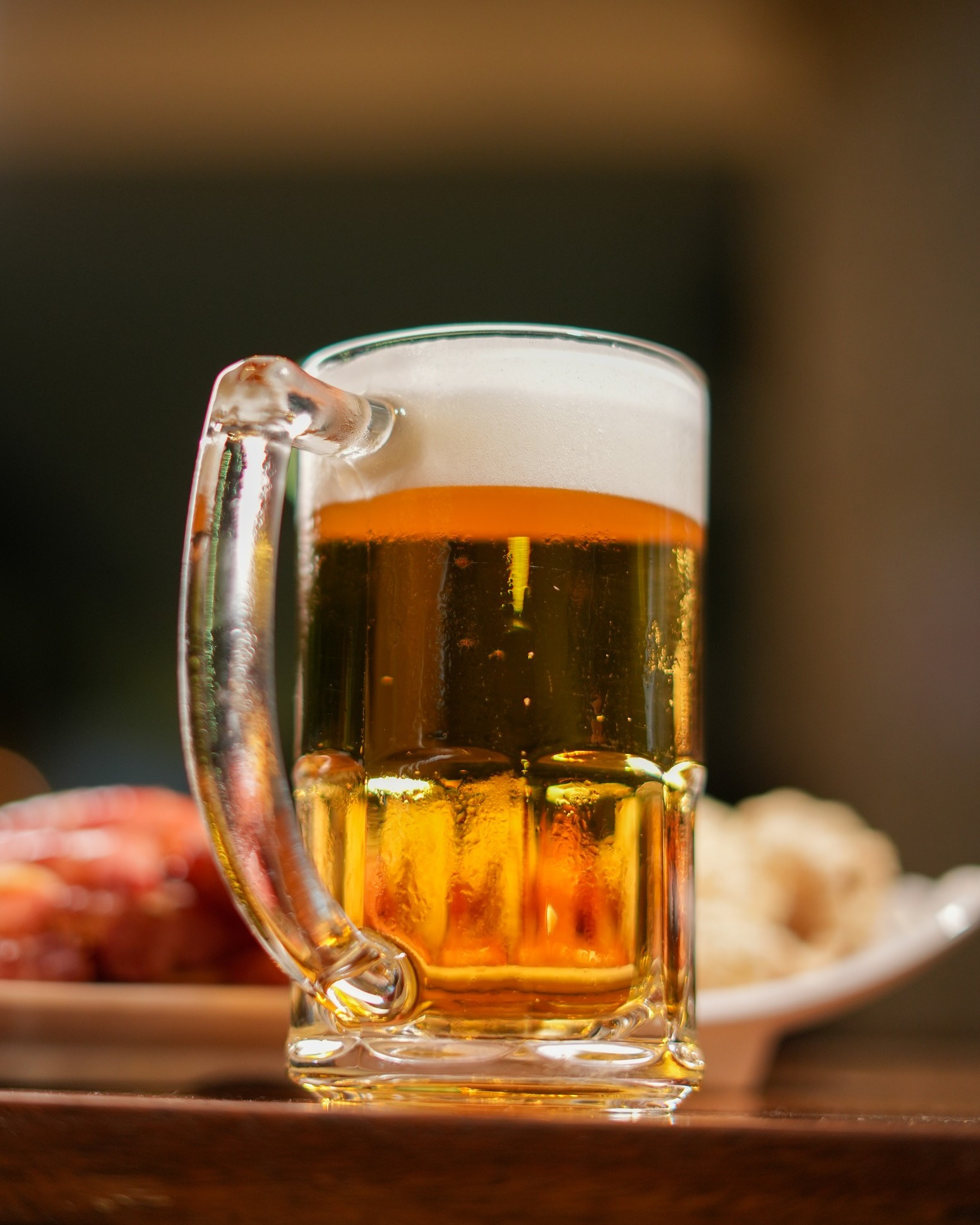 Caneca de chopp gelado com colarinho alto sobre balcão de madeira, com petiscos ao fundo